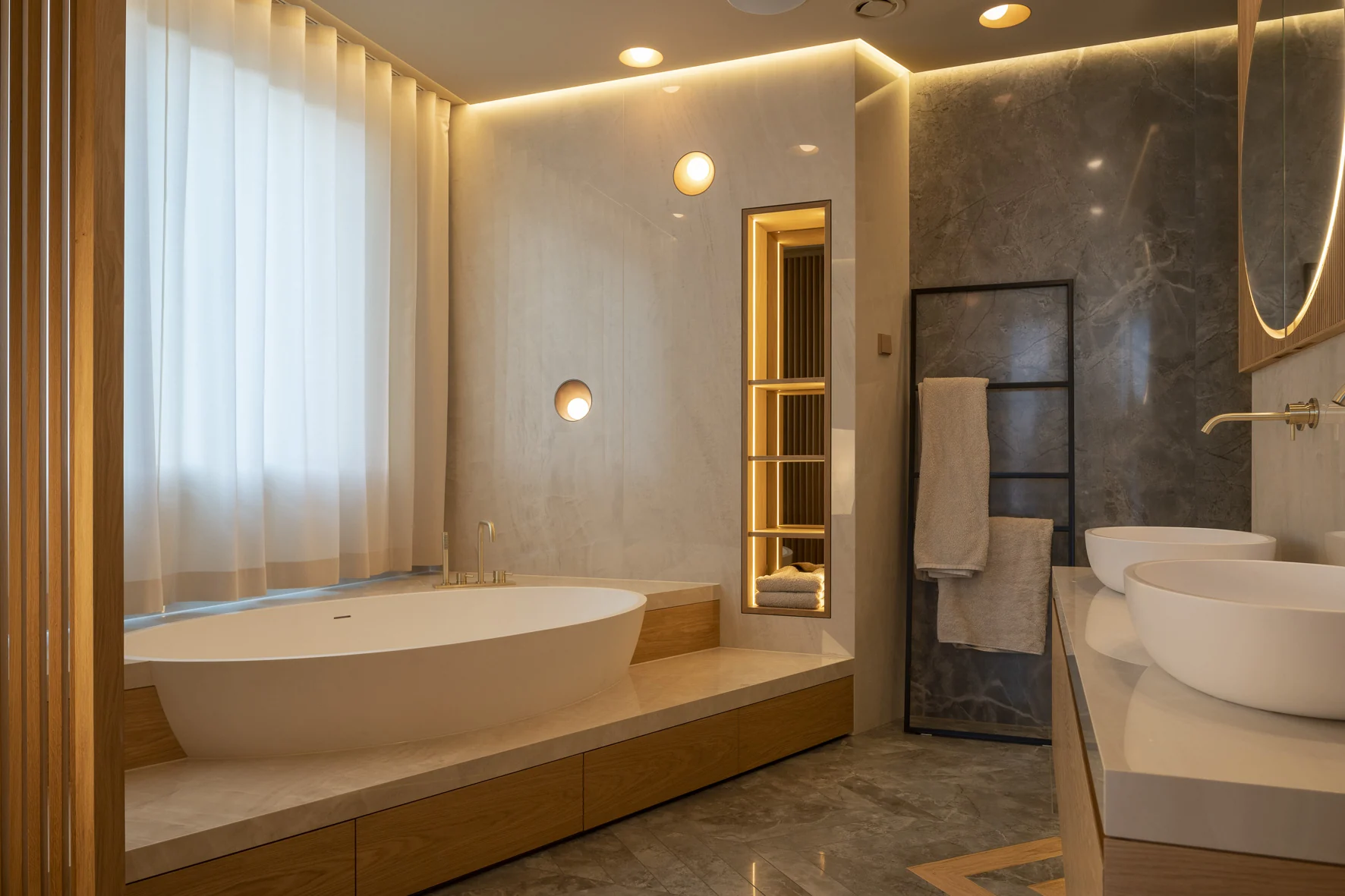 Masterbad mit freistehender Badewanne auf einem Natursteinpodest, maßgefertigten Holzeinbauten und warm beleuchteten Nischen