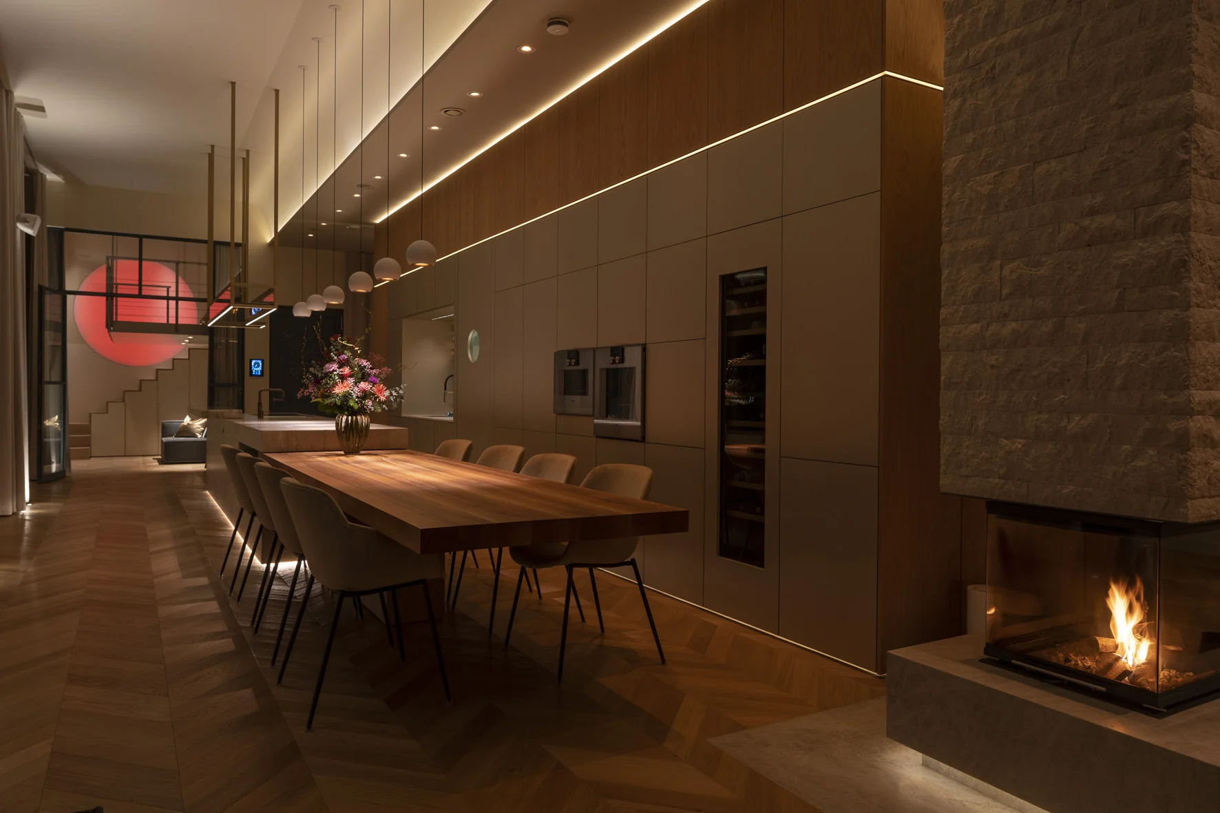 Wohn- und Essbereich bei Abendlicht mit langem Holztisch, Kamin und beleuchteter Treppe